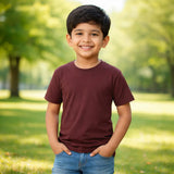 Young boy in a maroon plain suvin gold cotton t-shirt and blue jeans standing in a park with green trees and grass.