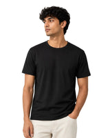 A man wearing a plain Supima cotton black short-sleeve crewneck t-shirt and off-white trousers, standing against a solid white background.