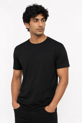 A man wearing a plain black short-sleeve crewneck t-shirt, standing against a light gray studio background with his hands in his pockets.