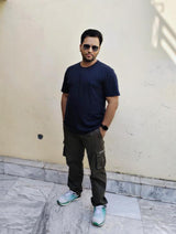 Man wearing a Supima Cotton  navy Blue t-shirt, cargo pants, and sunglasses standing against a light-colored wall.