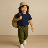 Child wearing a navy shirt, green pants, beige hat, and carrying a backpack on a beige background